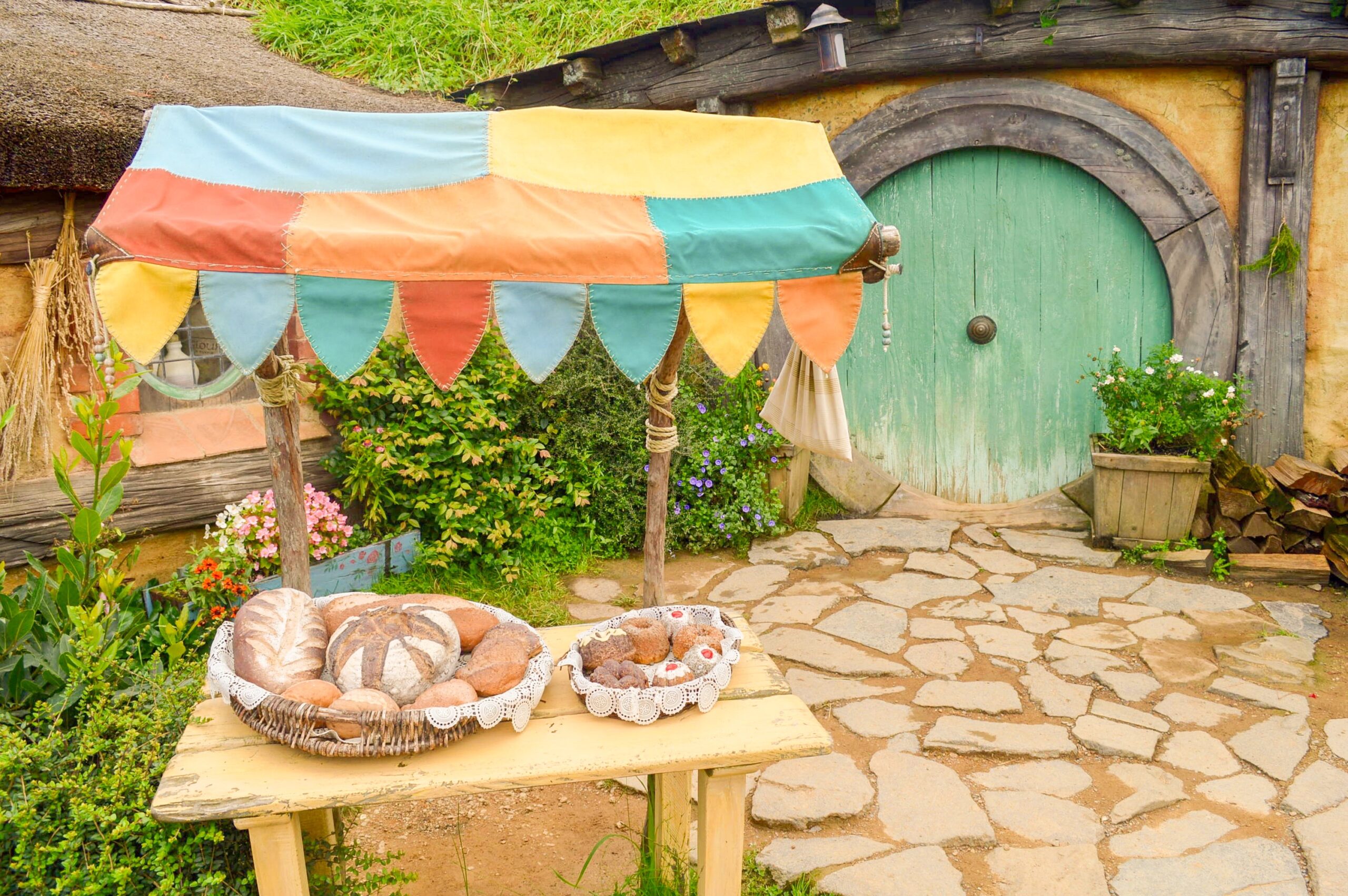 View of a baker's hobbit hole in Hobbiton with a small colorful cart of breads outside a green round door