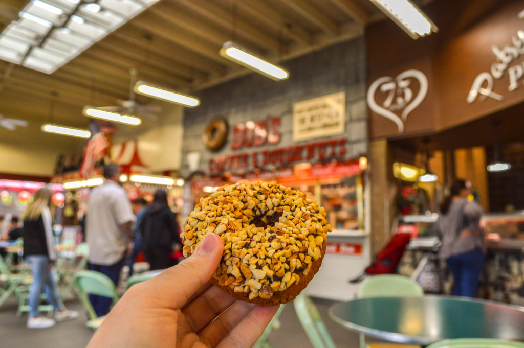holding a chocolate peanut covered donut with Bob's Donuts and Coffee in the Background at the Original Farmers Market in Los Angeles, California