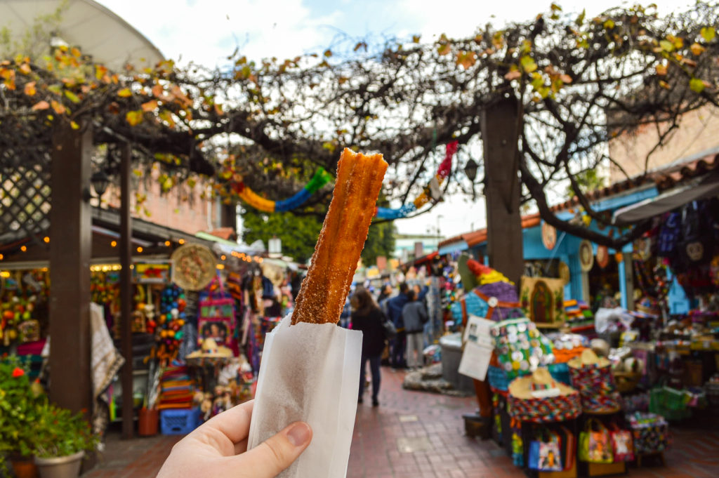 holding a churro up with a Mexican themed street in the background called Olvera Street, a highlight of a 5 day Los Angeles itinerary