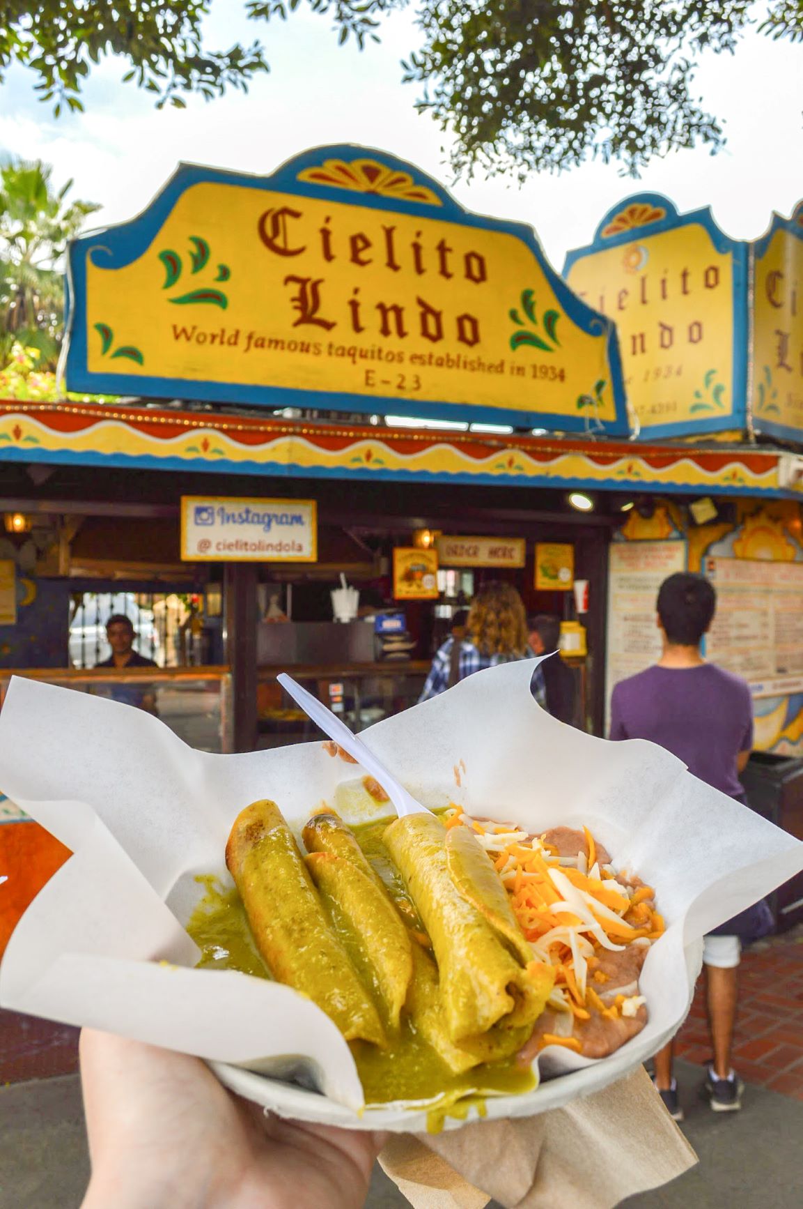Taquitos from Cielito Lindo on Olvera Street in a woman's hand outstretched with the food vendor booth behind