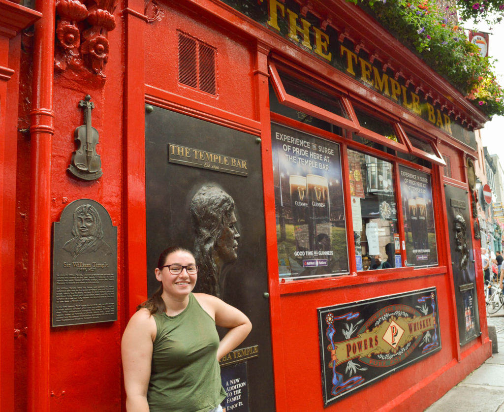 woman in an army green tank top standing in front of Temple Bar in Dublin Ireland, a bright red building