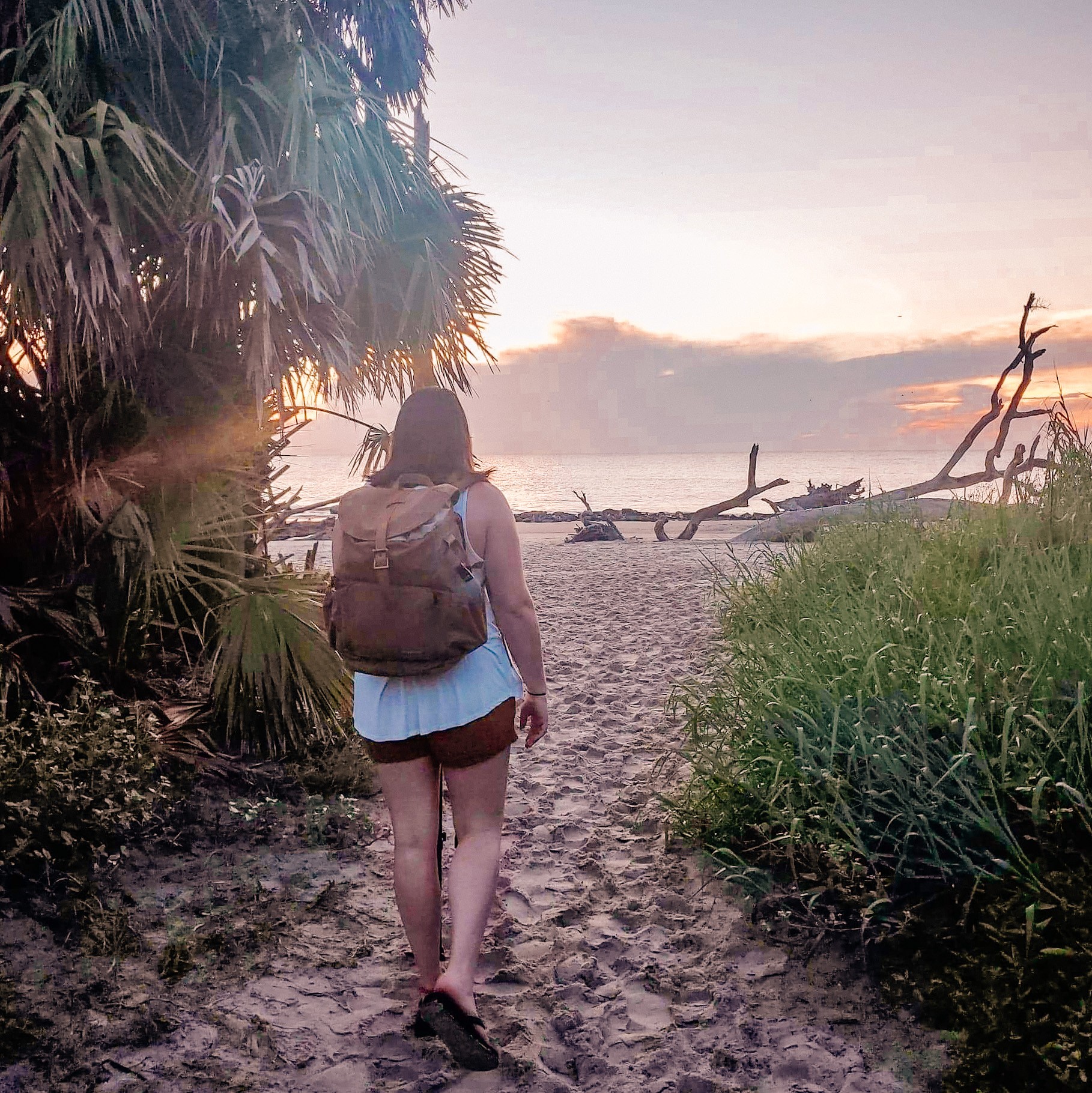 Woman walking on a path to the beach wearing a white tank top and khaki shorts and backpack