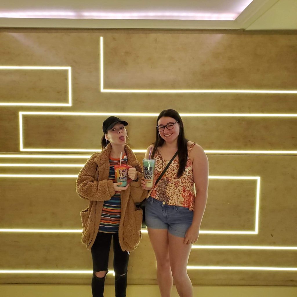 two women standing in front of a brightly lit geometric wall holding slushee beverages