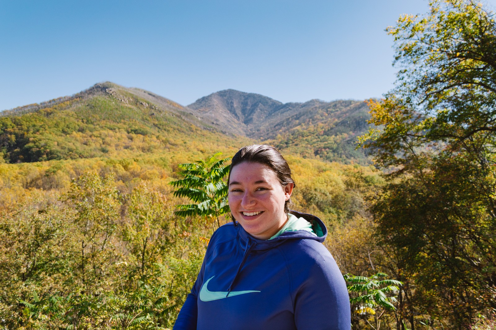 woman smiling at camera wearing a purple hoodie with yellow and orange fall foliage covered mountains behind her