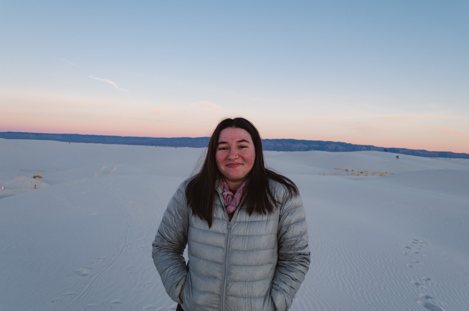 woman wearing puffer jacket grey in color with pink scarf tucked in and mid length hair down while looking at camera, standing on white sand dunes with pastel colored sunset glow in background