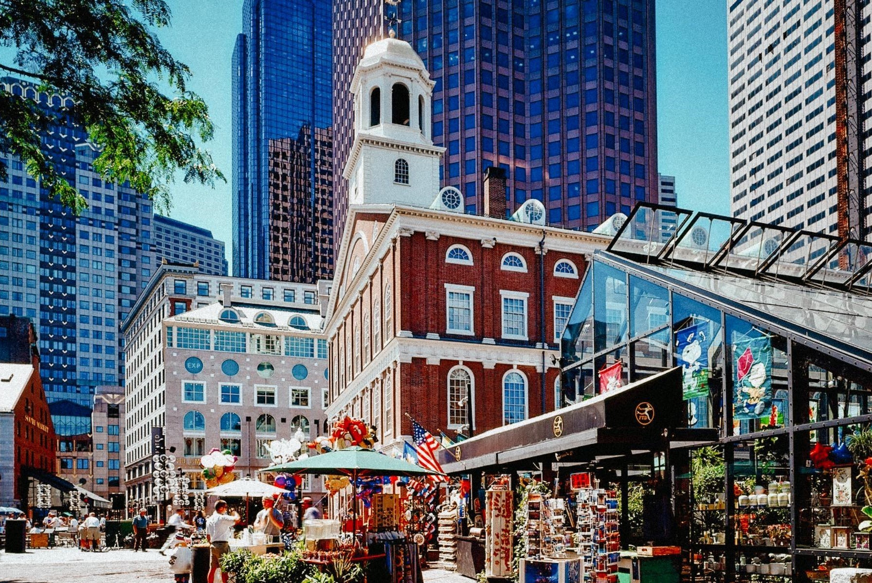 Collection of buildings of varying style and age in downtown Boston with skyscrapers in the background, an old historic building in the center with a tower (Faneuil Hall) and glass buildings on the sides
