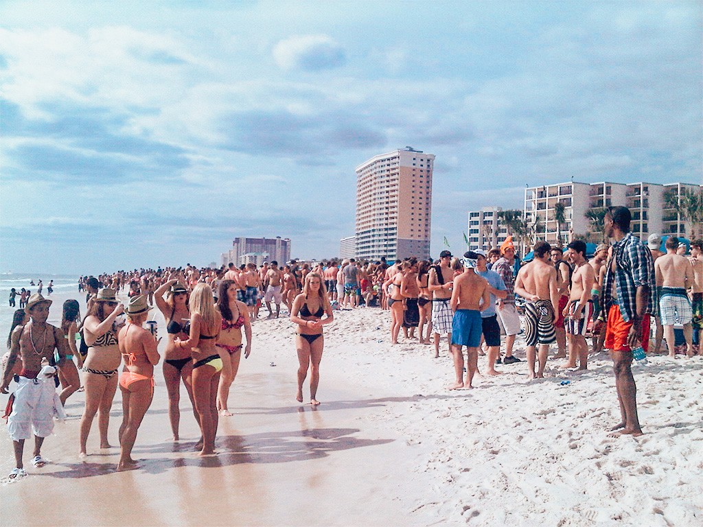 florida beach crowded with college age students and young people all in bathing suits partying, with white sand and partly cloudy skies