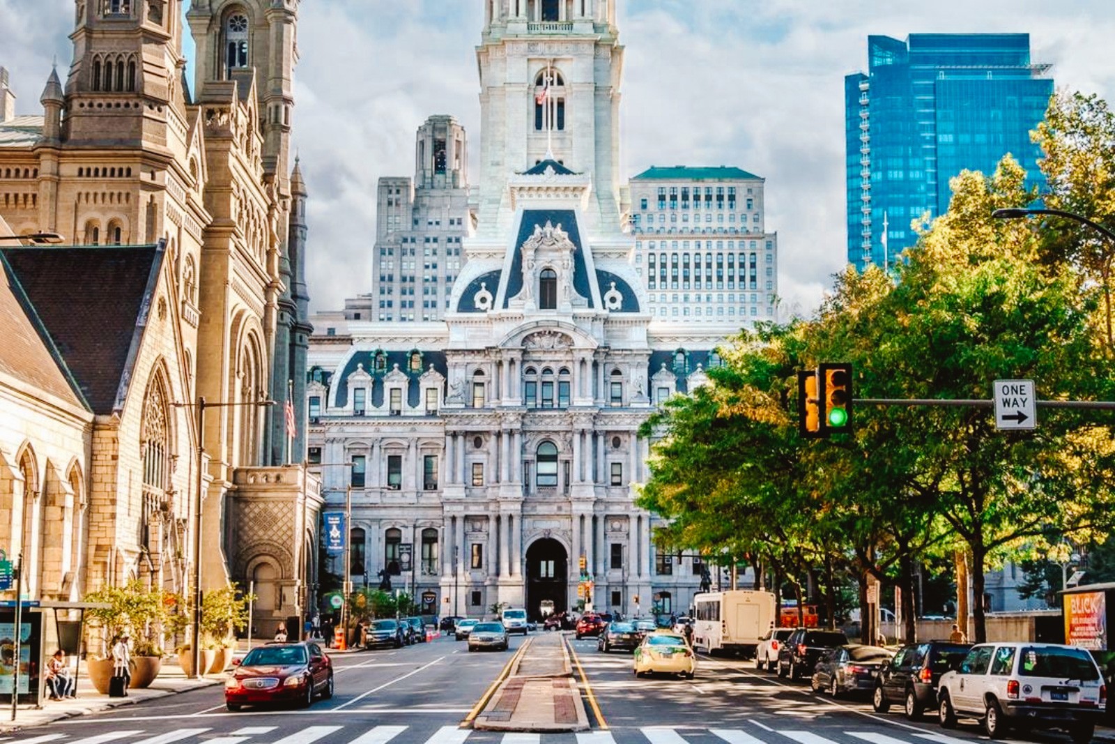 Beautiful buildings in Philadelphia on a sunny day with one white building centered with a street leading to it, trees on the right, and an old church with more beautiful historic architecture on the left