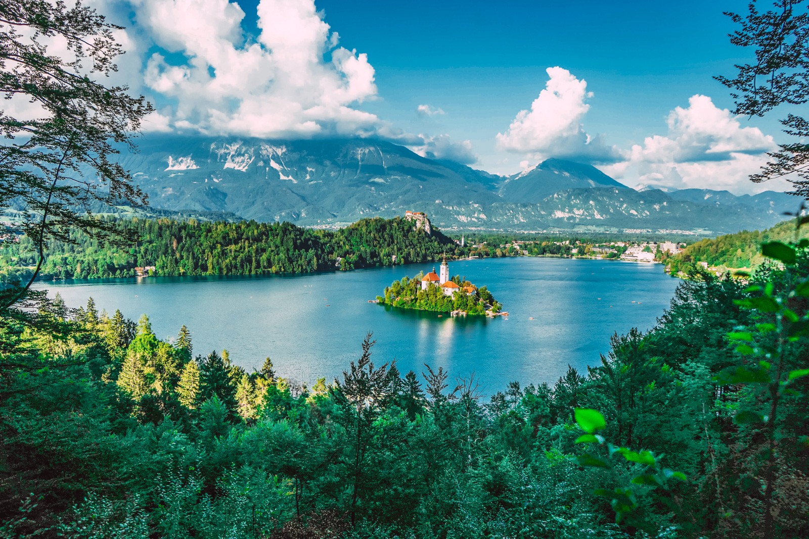 small island in the middle of a lake with a small castle like building on it with surrounding pine covered hills and mountains in the distance with partly cloudy sky