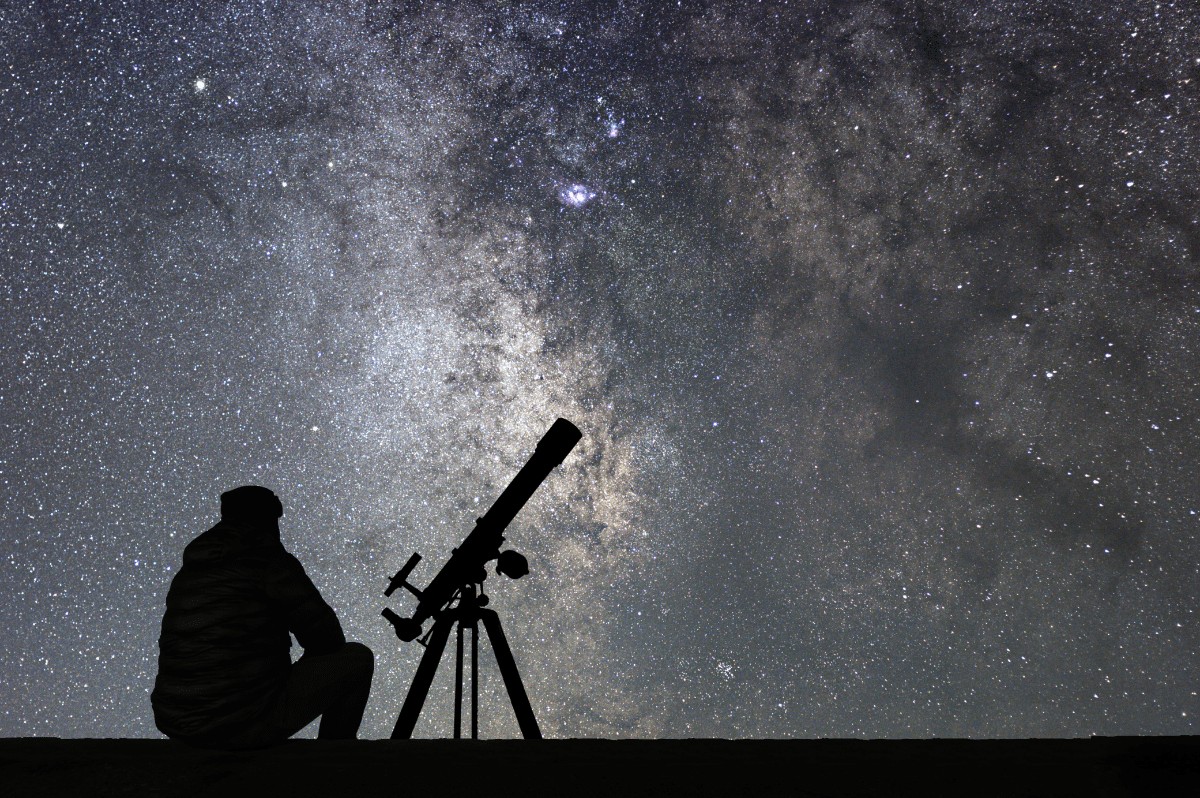 man stargazing with a telescope looking at the starry sky with galaxy clouds and nebulae in the background, man is silhoutted.