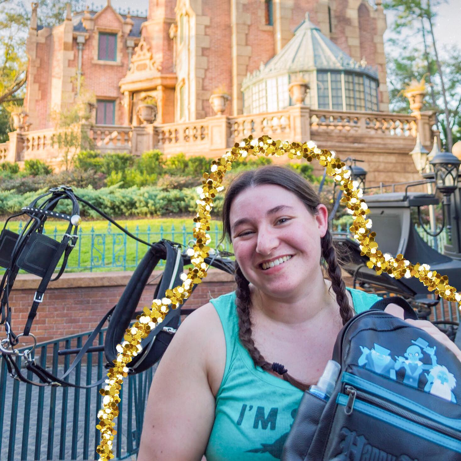 woman in braids smiling at the camera and holding a Haunted Mansion ride themed backpack in front of the ride itself