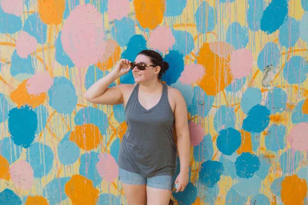 woman in grey tank top and jean shorts with sunglasses on looking to her right and holding the sunglasses while leaning on a mural wall with large dots of blue, orange, and pink colors