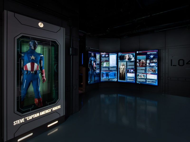 Captain America exhibit with uniform in a glass case on the left and images from the Marvel films in the distance on the right