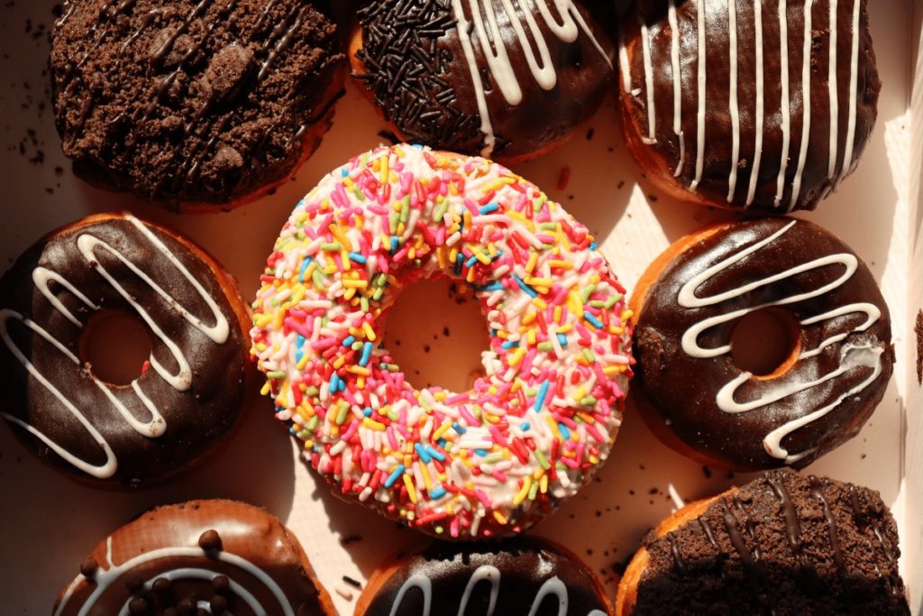 large donut with rainbow sprinkles in the center with 8 smaller chocolate donuts surrounding in a donut box