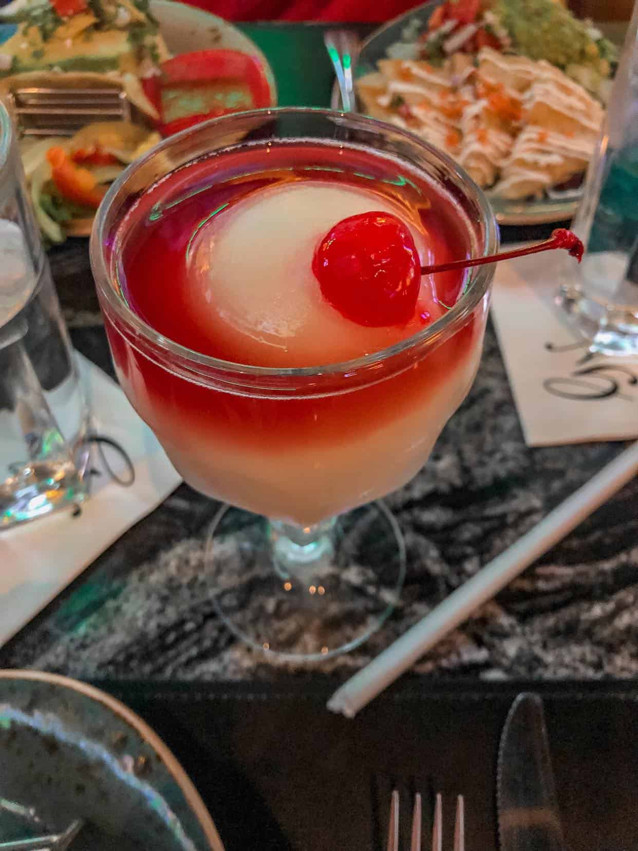 frozen margarita with red syrup topping and a cherry on top with mexican food on the table surrounding
