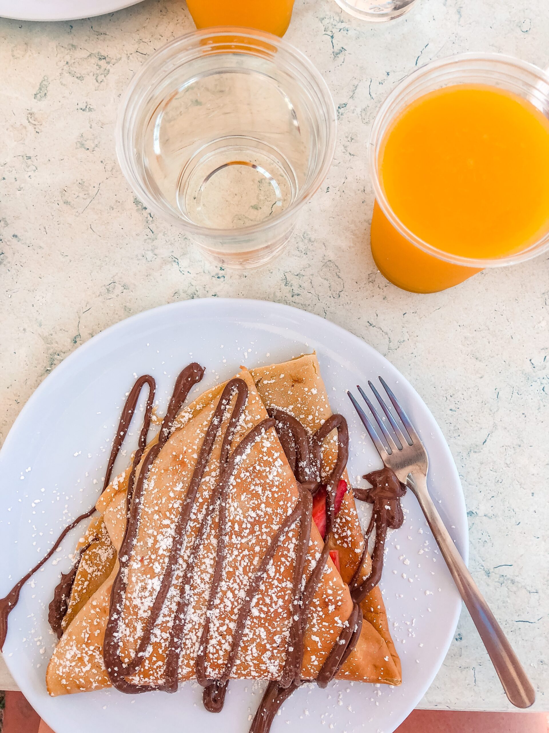 view of breakfast meal from above in a flat lay image of a nutella and strawberry crepe, glass of orange juice and water to the top right