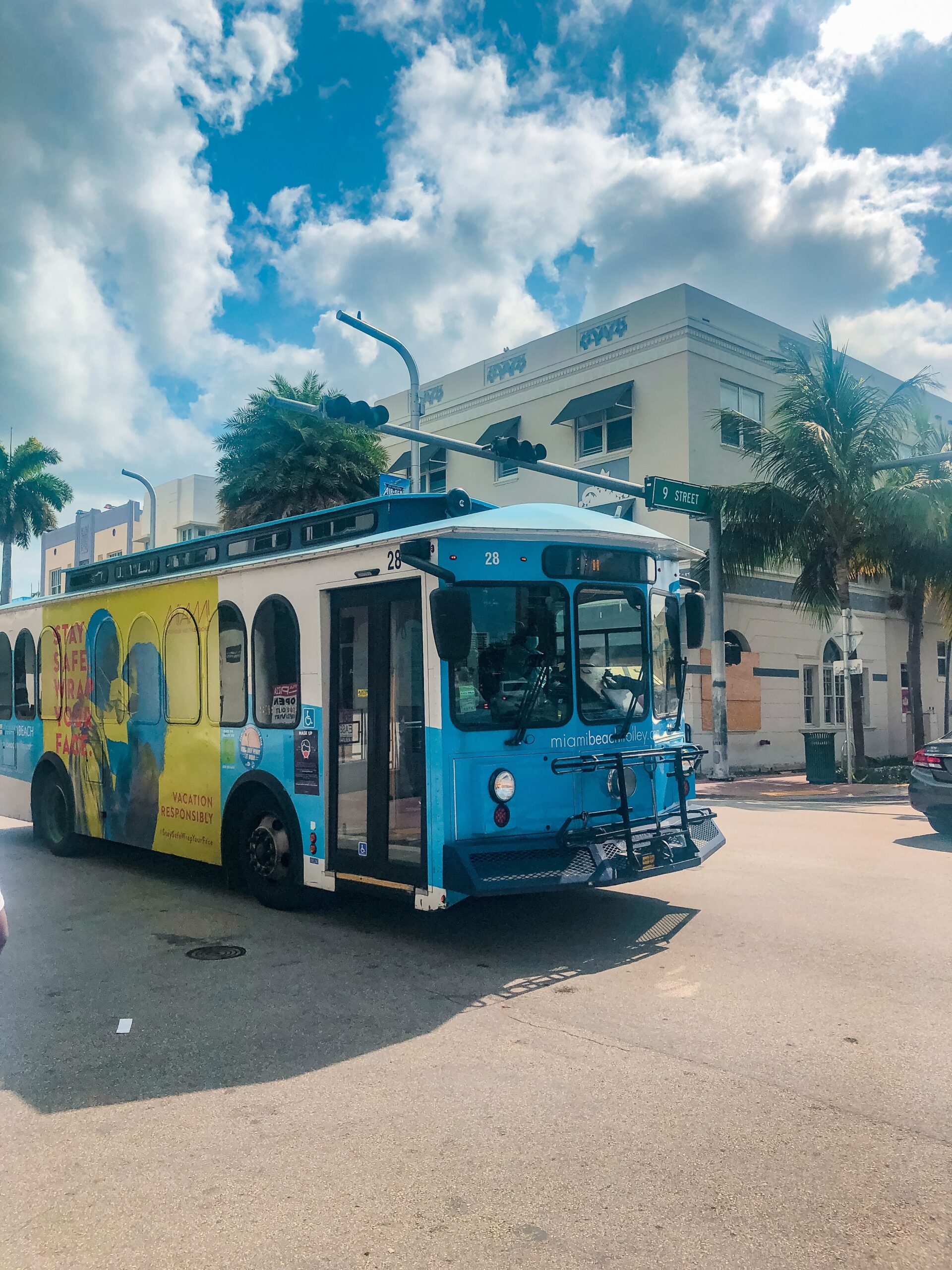 Blue trolley bus driving independently on a street in Miami Beach Florida with partly cloudy skies and a cream colored building in the background with a few palm trees scattered along the street