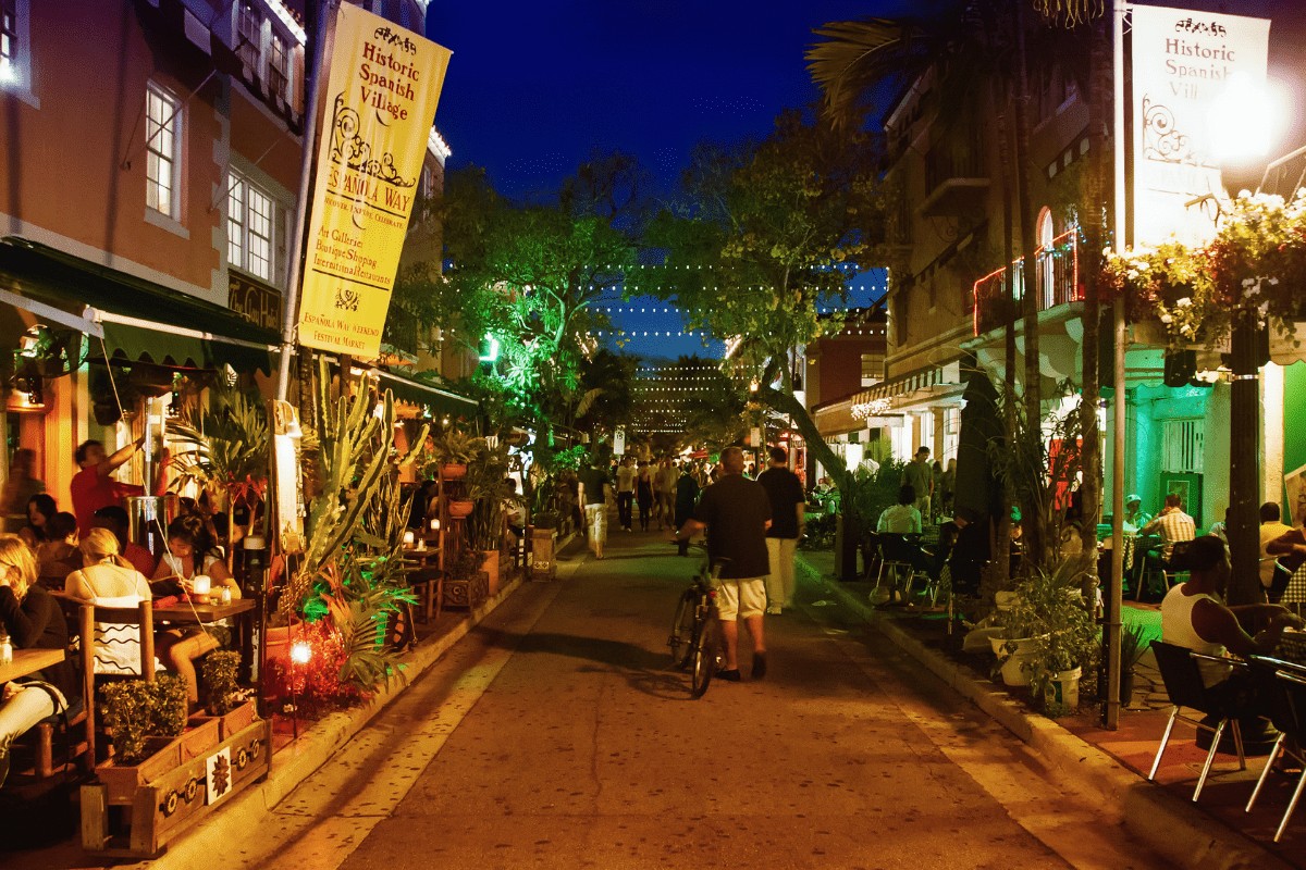 view of a pedestrian street at night with restaurants on both sides that have large outdoor dining areas and a yellow sign that reads Espanola Way
