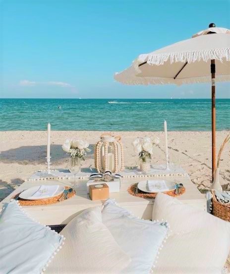 professional picnic display on the beach with a small table, lots of pillows to sit on, two place settings and an umbrella with turquoise ocean in the background