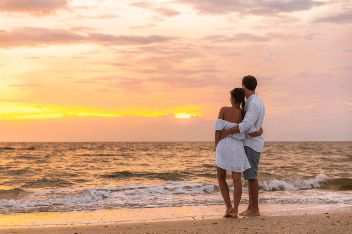 man and woman couple dressed in white looking away from camera at ocean sunset with orange, pink, and yellow colors