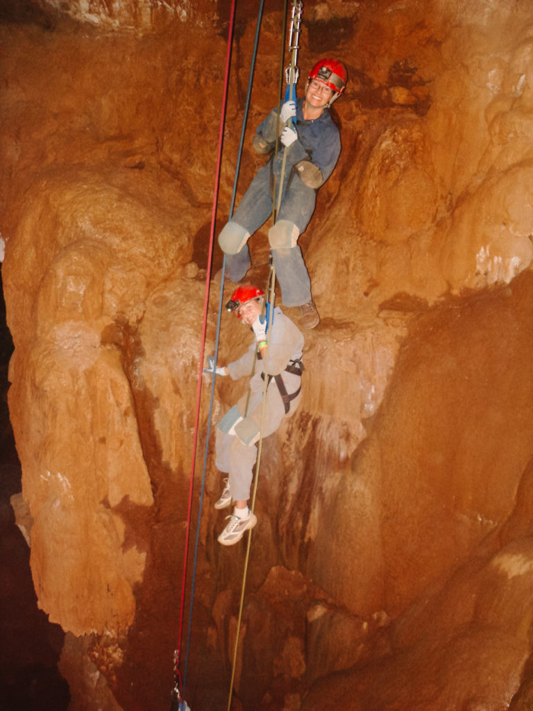 a mom and daughter descending a cave wall in spelunking gear like helmets and jumpsuits