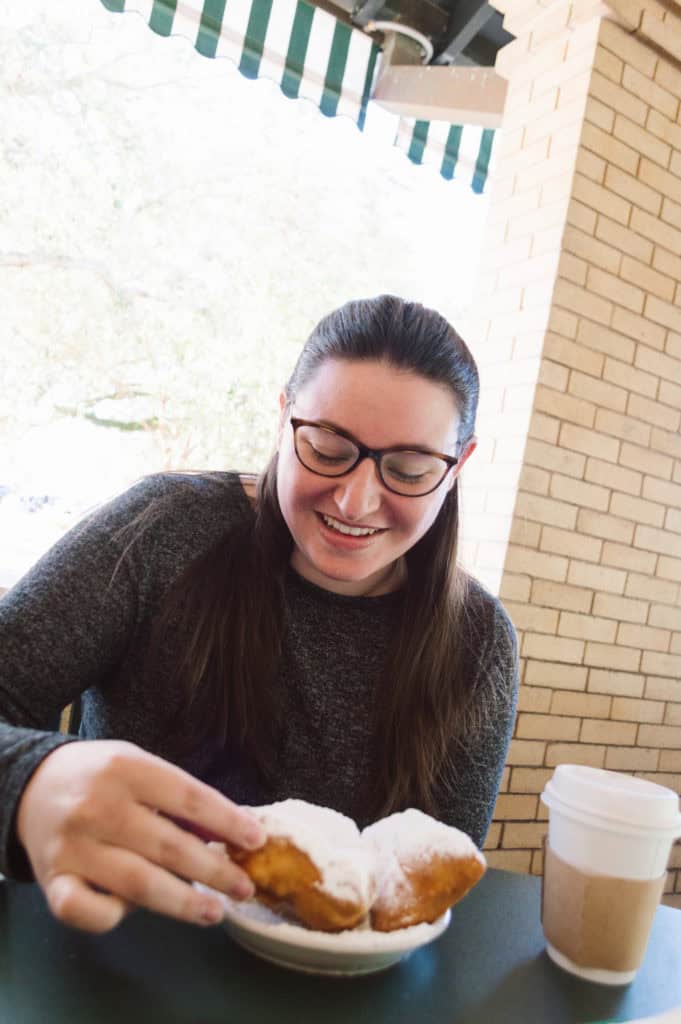 woman with long brunette hair and glasses about to eat beignets with a coffee to her left