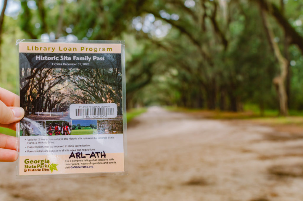 holding Historic Site Family Pass curtesy of the Library Loan Program in Georgia with Wormsloe Historic Site in the background with spanish moss covered trees arched over the dirt road