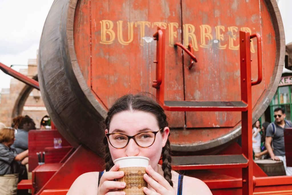 woman sipping hot butterbeer from a to-go cup with a red butterbeer barrel behind her