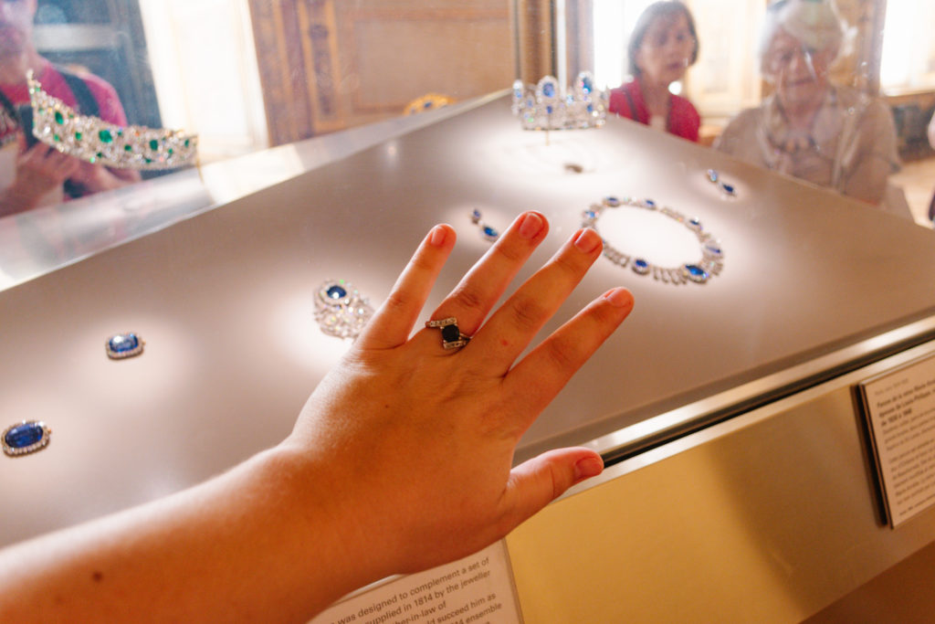 woman's hand extended with a sapphire engagement ring with other sapphire jewelry in a case behind her hand
