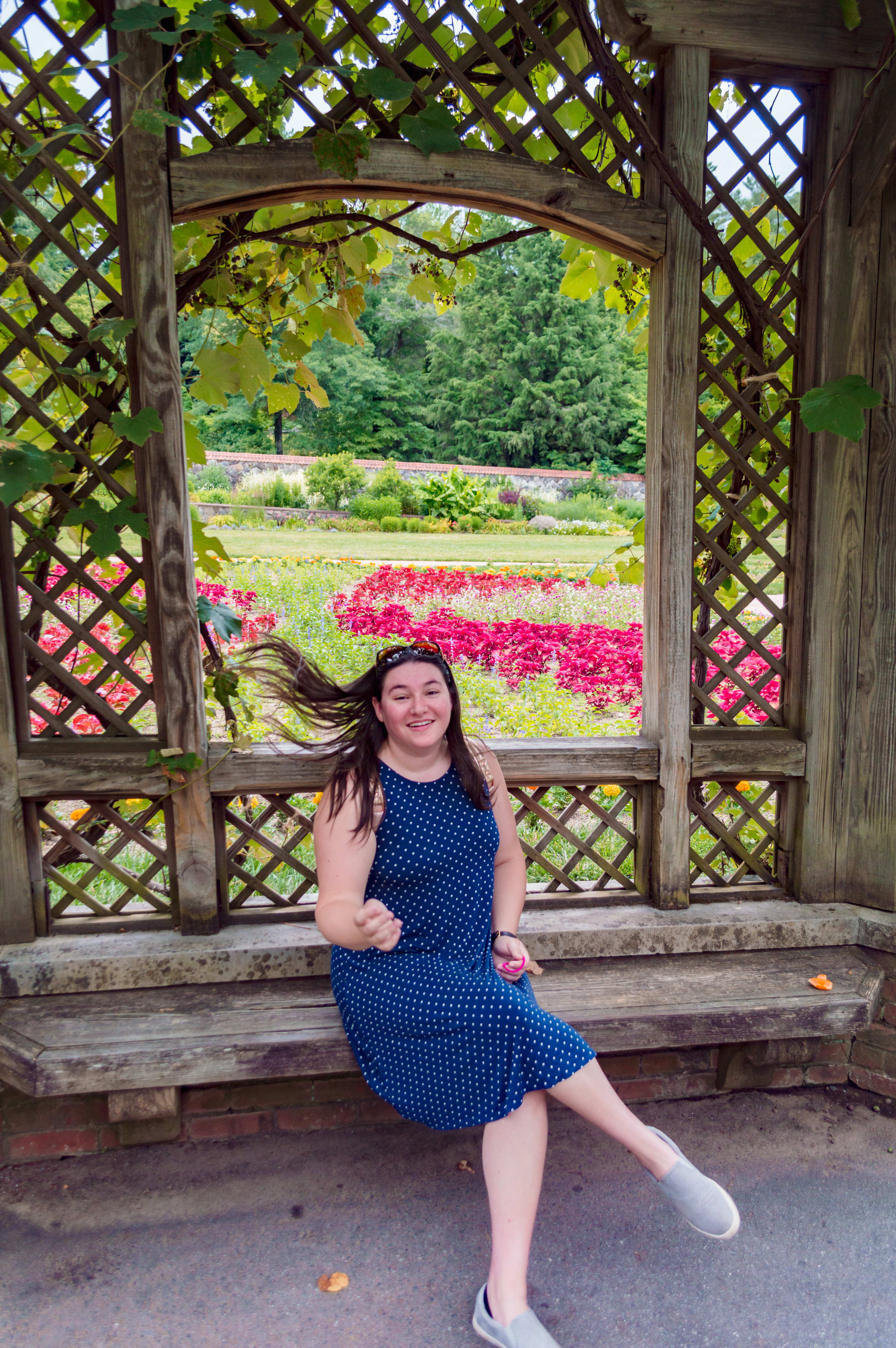 woman in blue dress flipping her hair while seated on a bench under a covered terrace in a rose garden