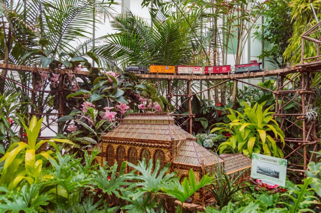 indoor conservatory with a lot of vegetation surrounding a model building with a train track elevated and a model train car passing by.