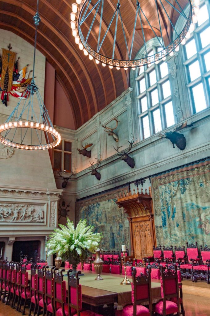 Seven story tall dining room with arched wood ceiling large circular chandeliers and dining room table with red chairs.