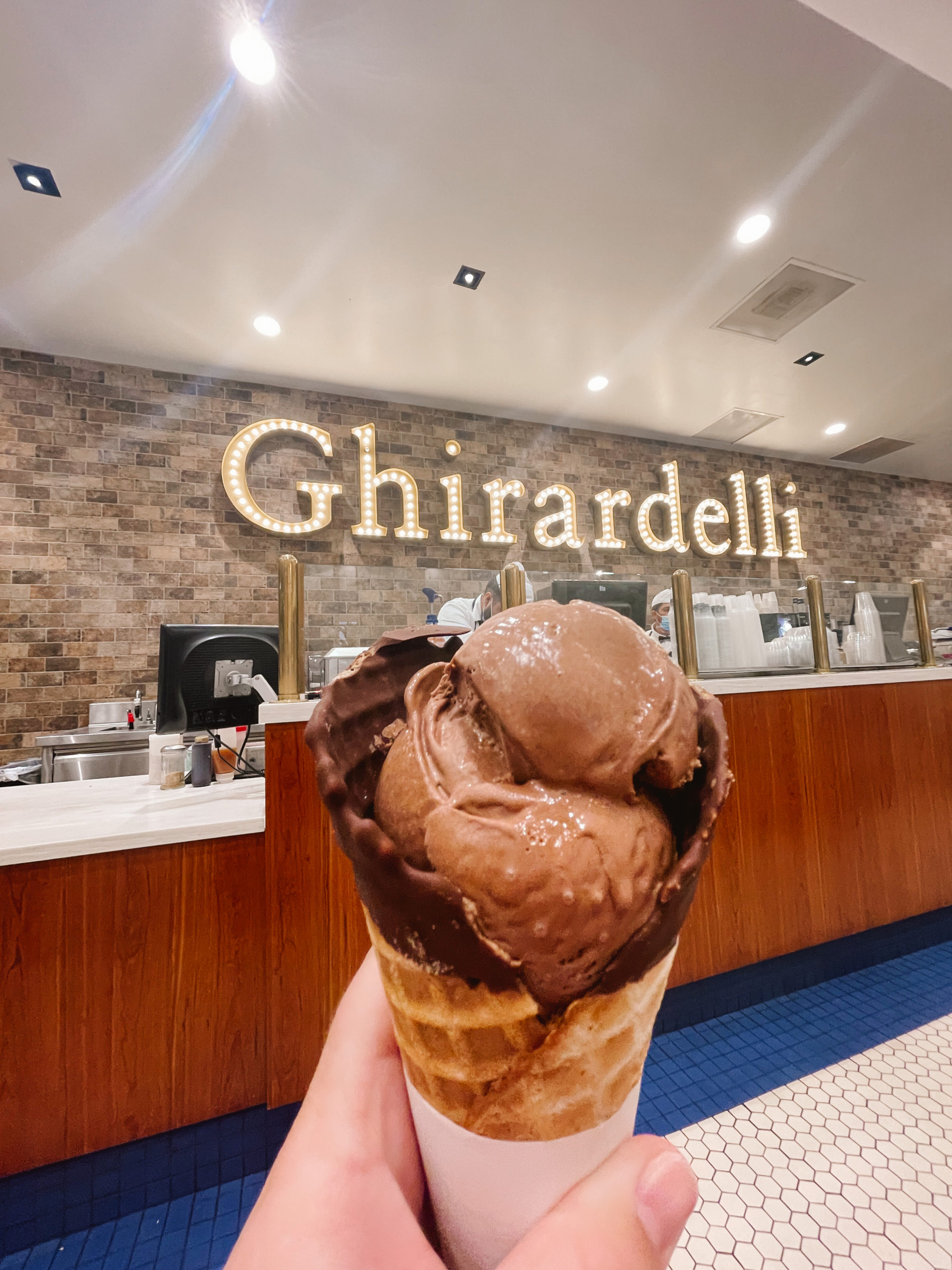 hand outstretched from camera holding chocolate ice cream cone with Ghirardelli neon sign on brick wall in background