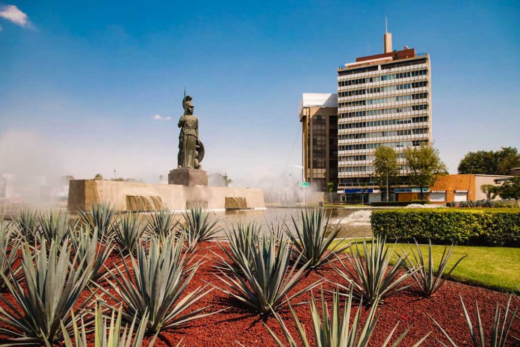 City park of Guadalajara, Mexico with statue fountain in center, desert plants surrounding, and modern building in the background on a sunny day