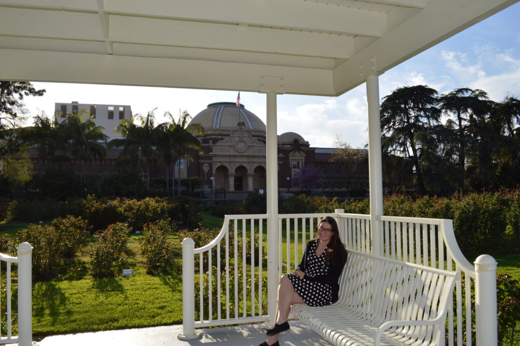 woman sitting on a white bench on a covered platform with a museum behind her and rose garden surrounding