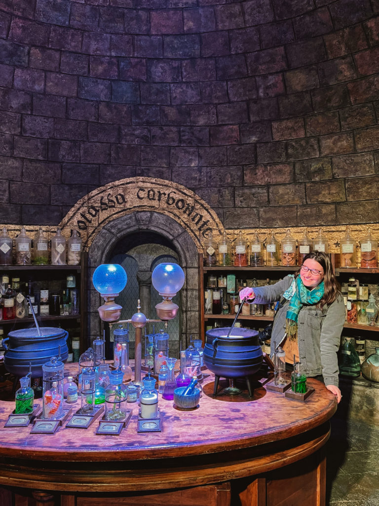 woman in grey jean jacket and bright turquoise scarf stirring a potion in a potions classroom from Harry Potter