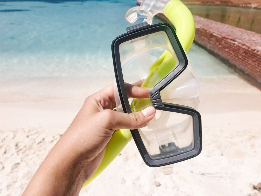 woman's hand outstretched from camera holding swim mask and snorkel with ocean in the background