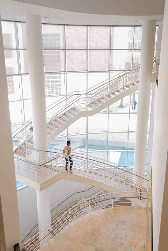 man walking down white stair case with bright light coming through floor to ceiling windows behind him.
