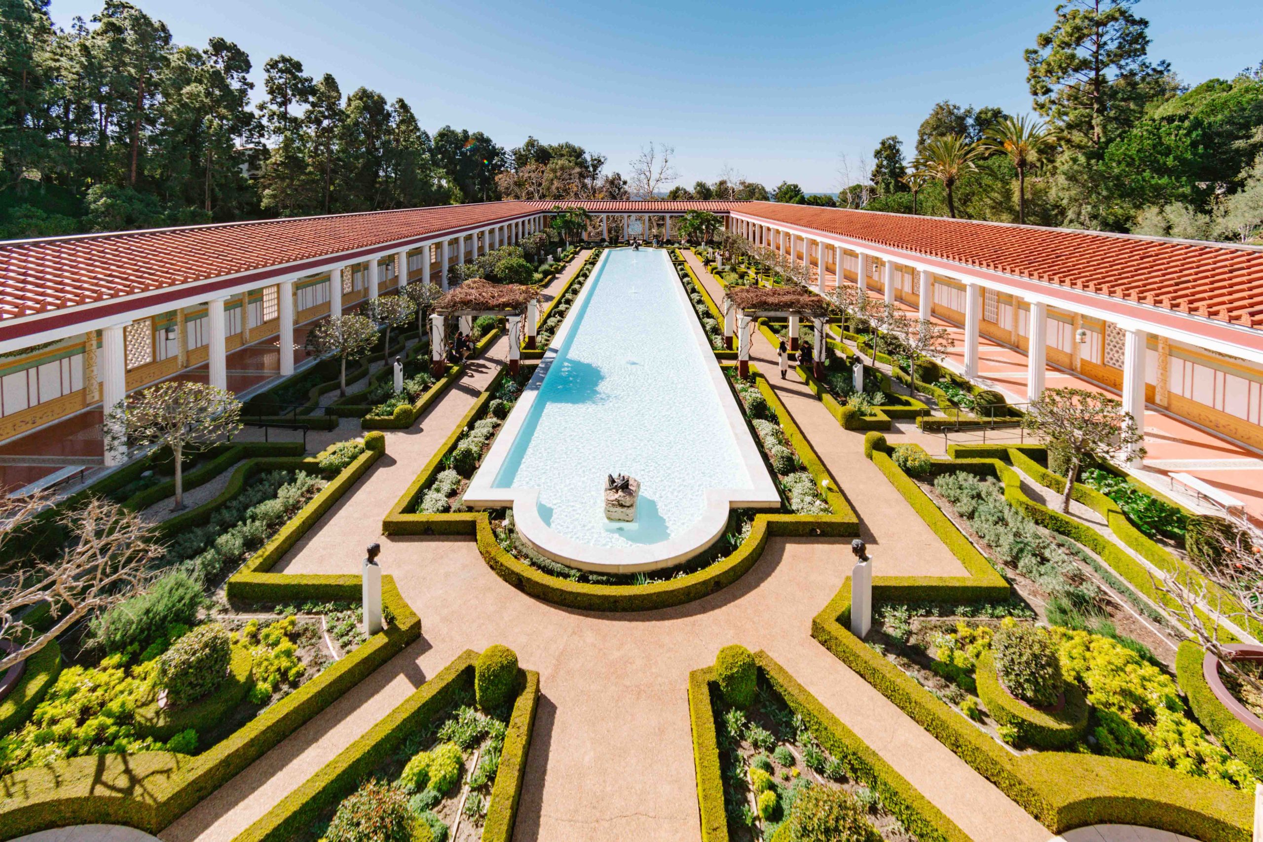 wide angle view of the Getty Villa in Pacific Palisades with long Roman pool in the center, topiaries surrounding and long tiled hallways adjacent.