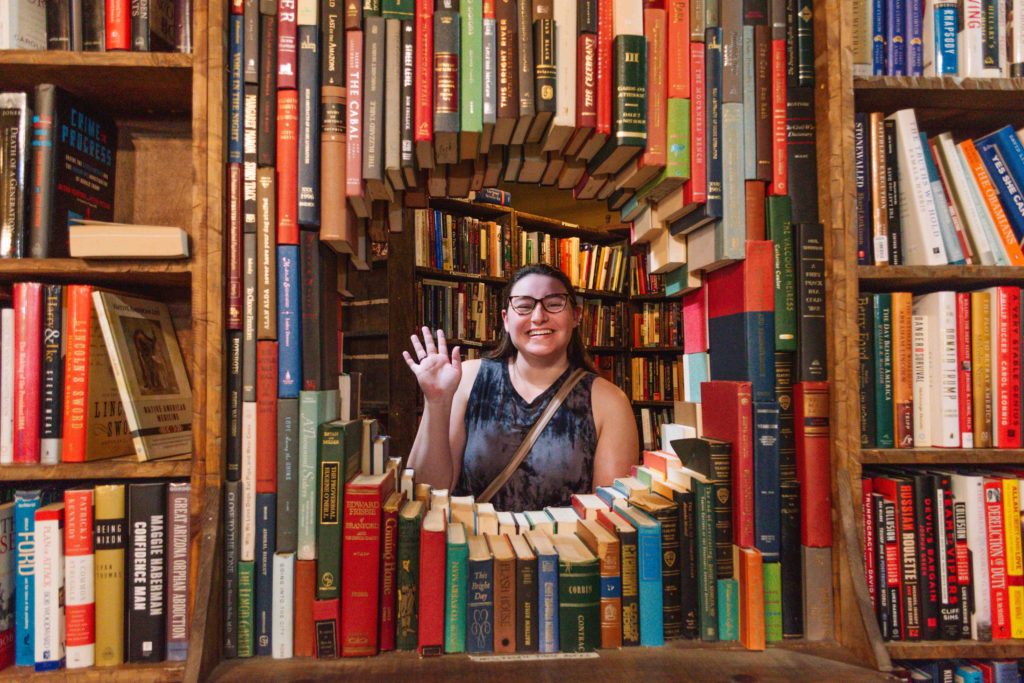 woman waving from behind a window made from books in a book store