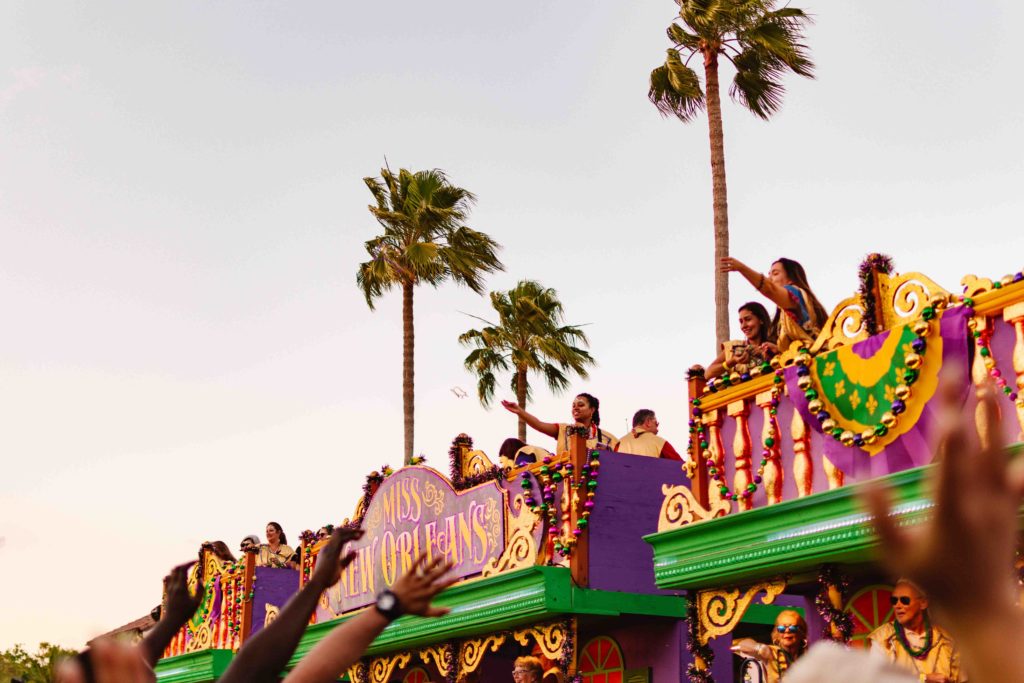 people throwing beaded necklaces off parade floats to a crowd below with palm trees in the background