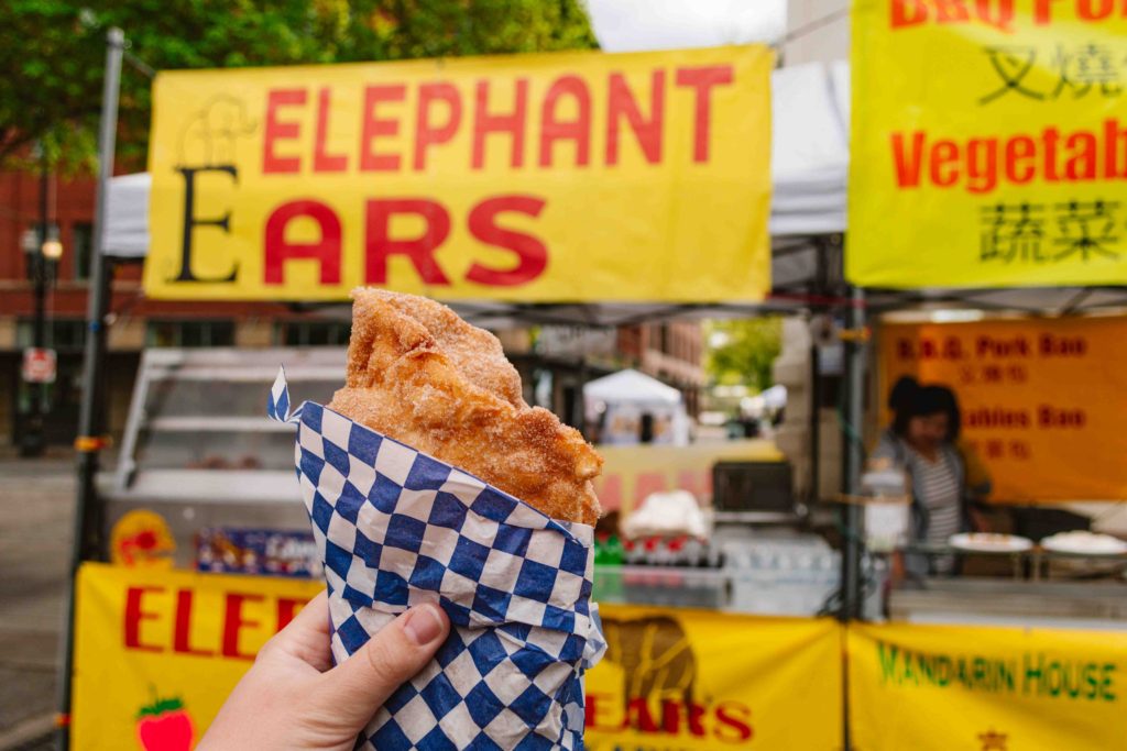 outstretched hand holding "elephant ear" snack, a cinnamon sugar covered fried dough with vendor stall in the background