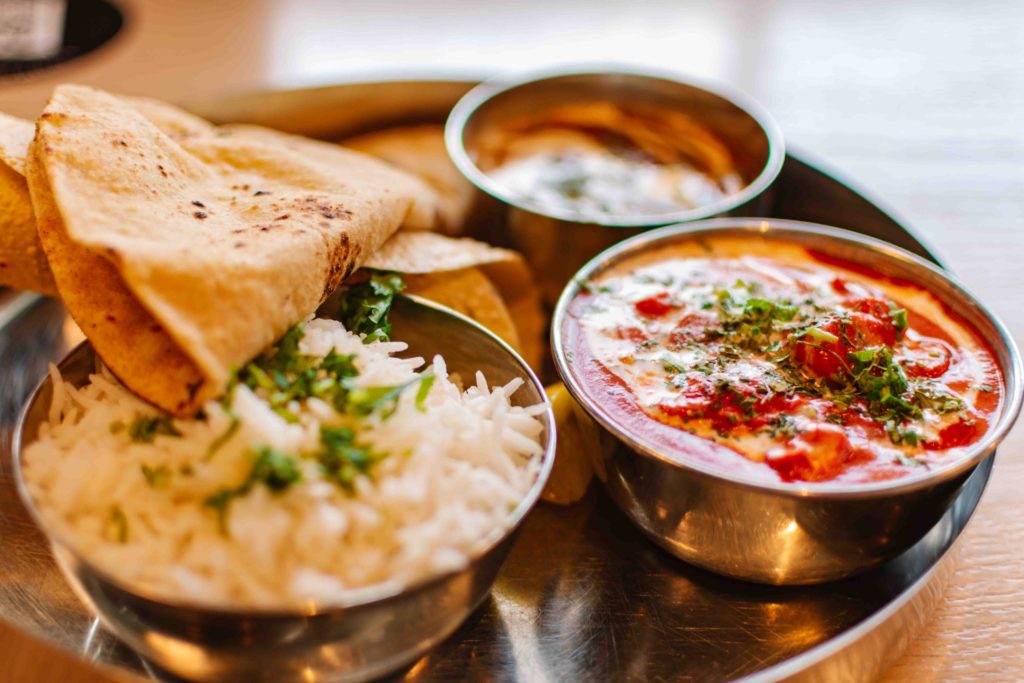 Brushed steel plate of small bowls of Indian food dishes including chicken tikka masala, basmati rice, daal, and roti