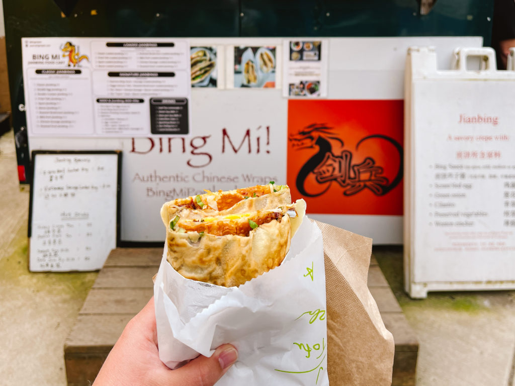 Jianbing in a woman's outstretched hand with the food cart in the back that made the Chinese savory pancake
