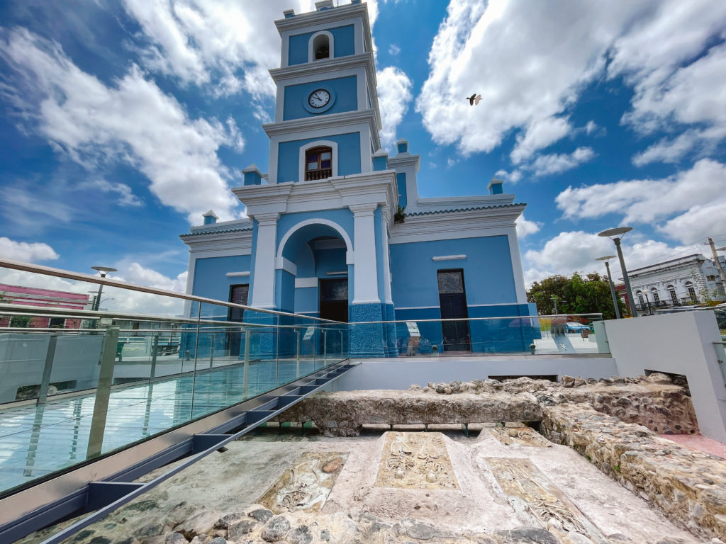 Blue religious cathedral with a class bridge to the entrance and replica skeleton burials at the foot of the church under the glass bridge