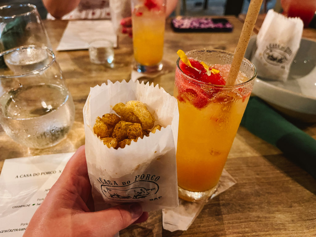 outstretched hand holding a bag of pork skins with tropical drink to the right