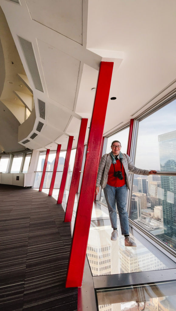 Woman standing on a glass floor atop Calgary Tower in Alberta, Canada wearing a red shirt, grey jacket, and blue jeans