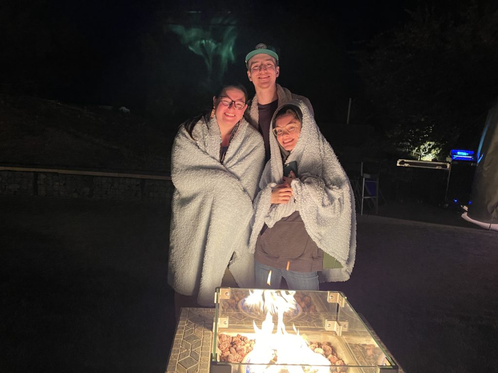 two women and a tall man standing by a firepit bundled in blankets