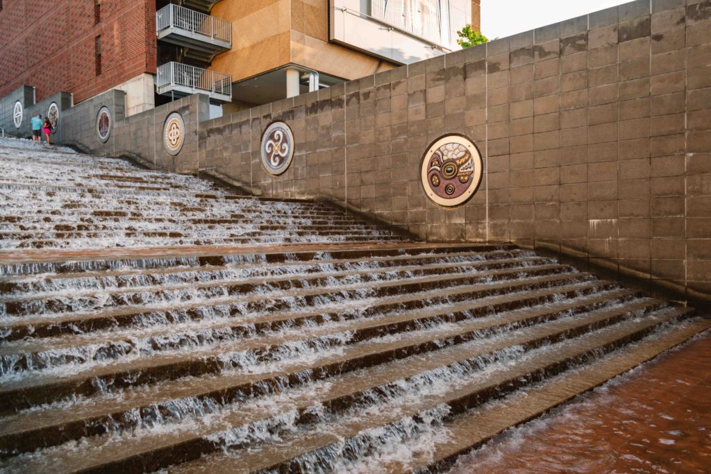 Cascading water falling over concrete steps with a wall adjacent that has symbols of seven Cherokee clans