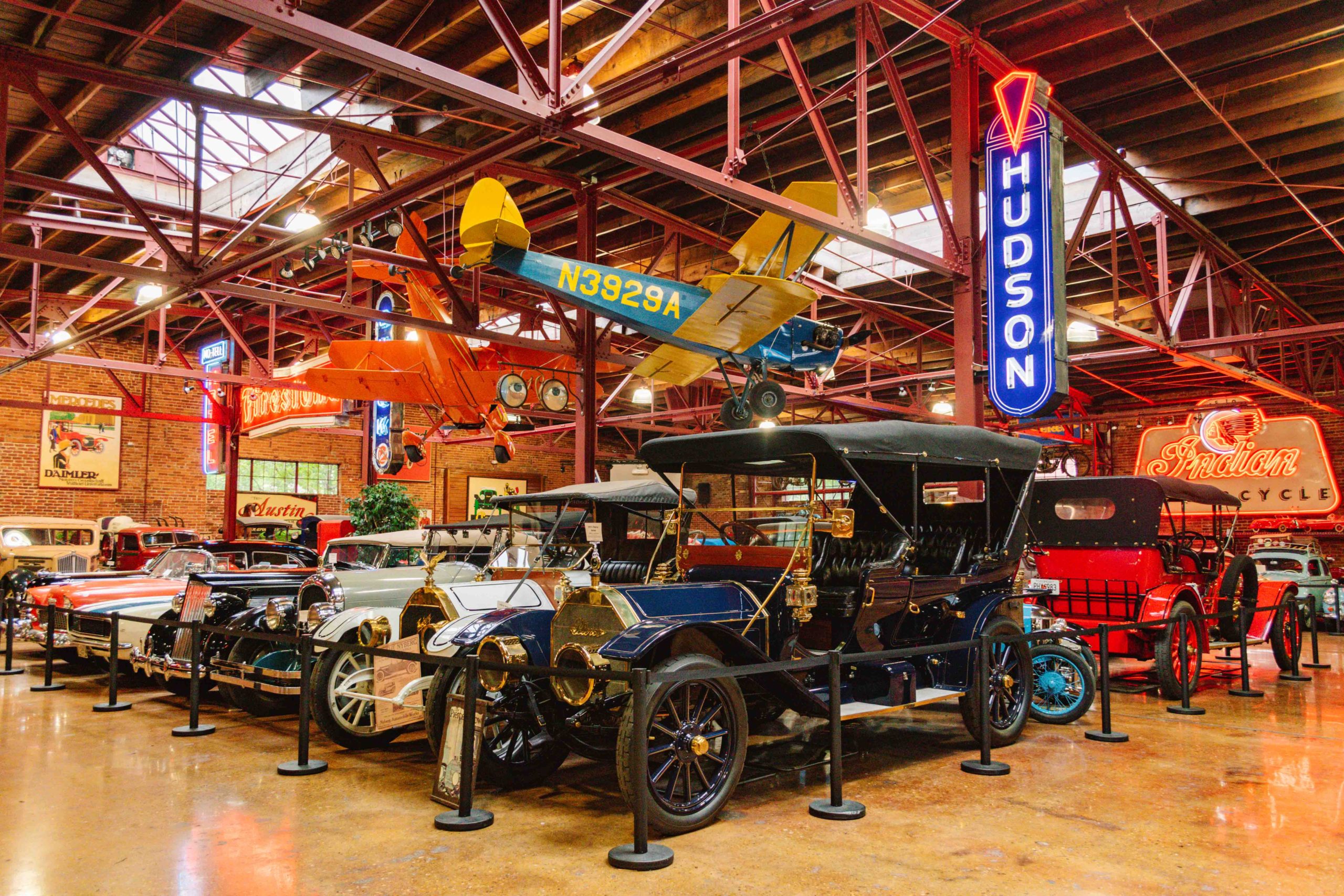 Museum warehouse full of vintage cars and neon signs plus 3 airplanes suspended from the ceiling.